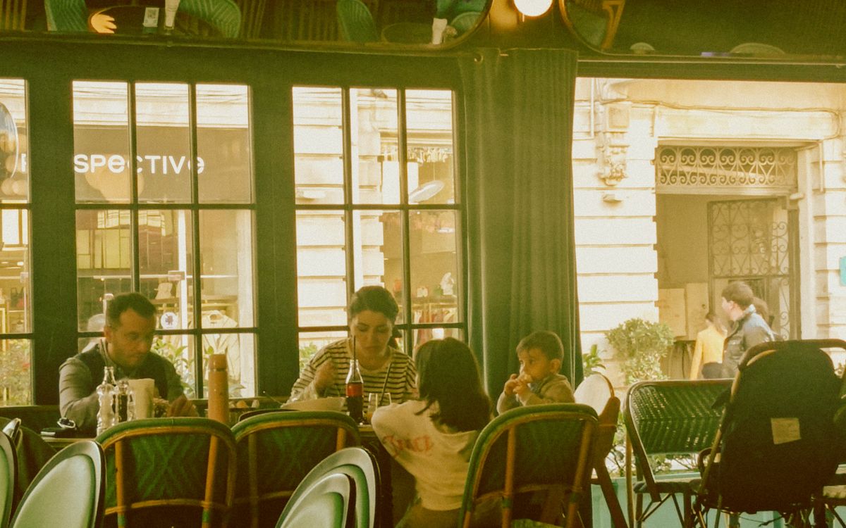 Família com dois filhos almoçando à mesa de um café ao lado de uma grande janela, em um dia claro