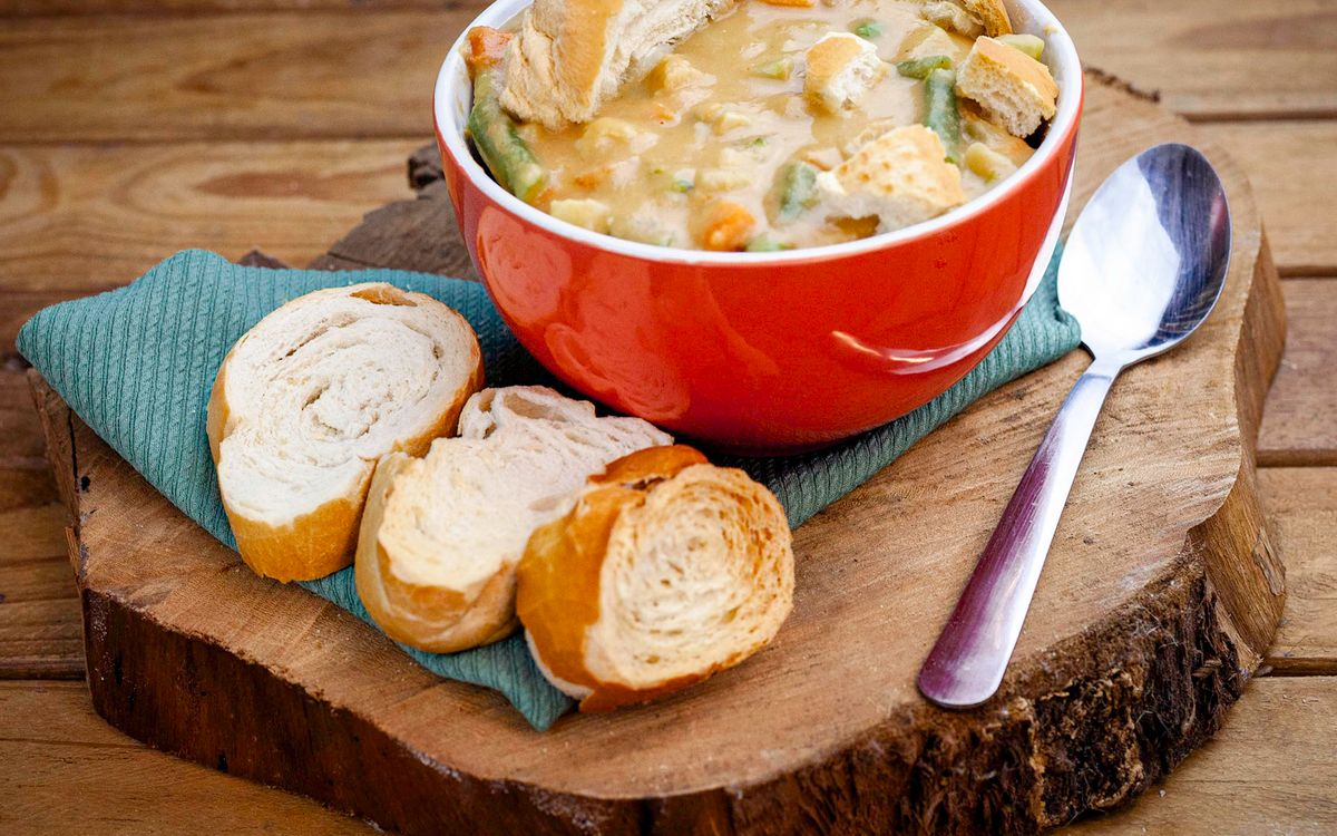Tigela vermelha com caldo cremoso de legumes ao lado de fatias de pão sobre uma tábua de madeira rústica