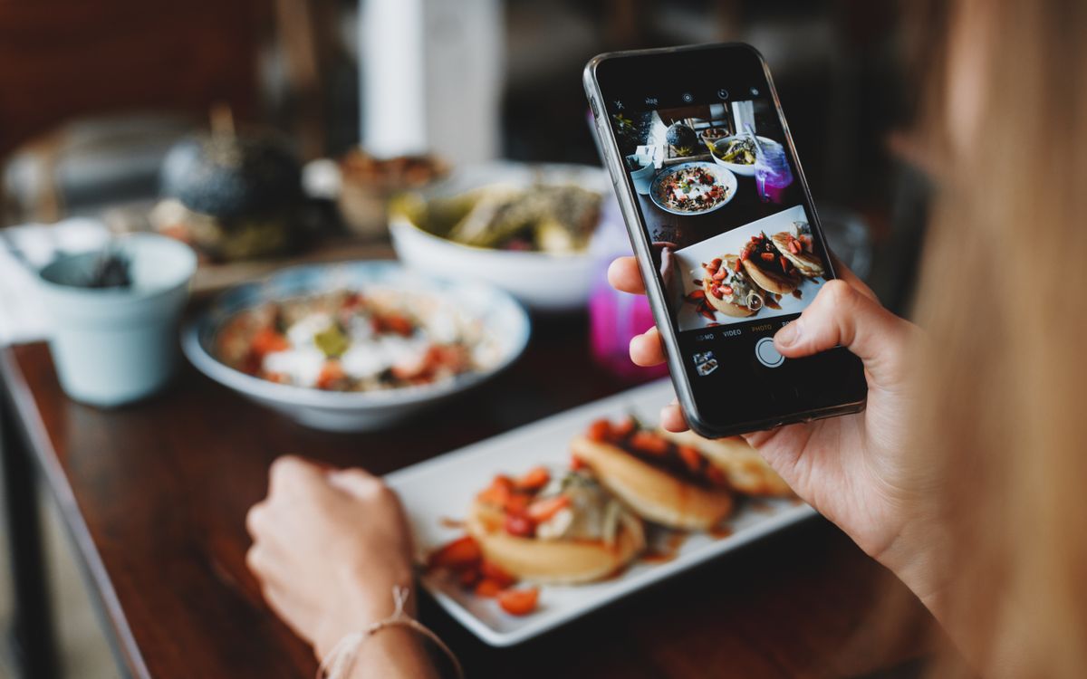 Mão segurando um celular que fotografa um prato de panquecas com tomate sobre a mesa de madeira de um restaurante
